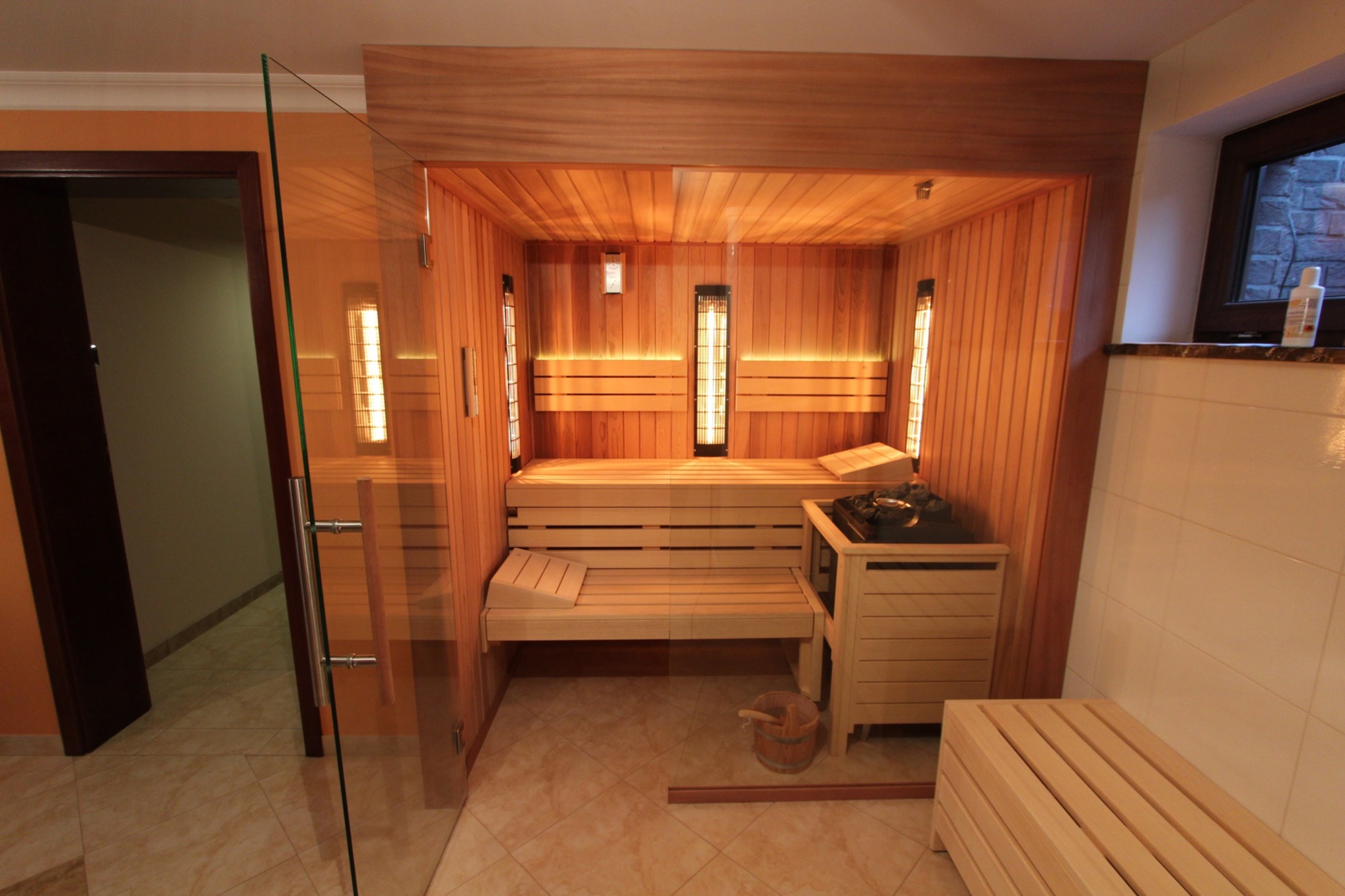 Wnętrze sauny z drewnianymi ławkami, piecem na kamienie i przeszklonymi drzwiami, oświetlonej lampami grzewczymi, widok z zewnątrz przez otwarte drzwi.