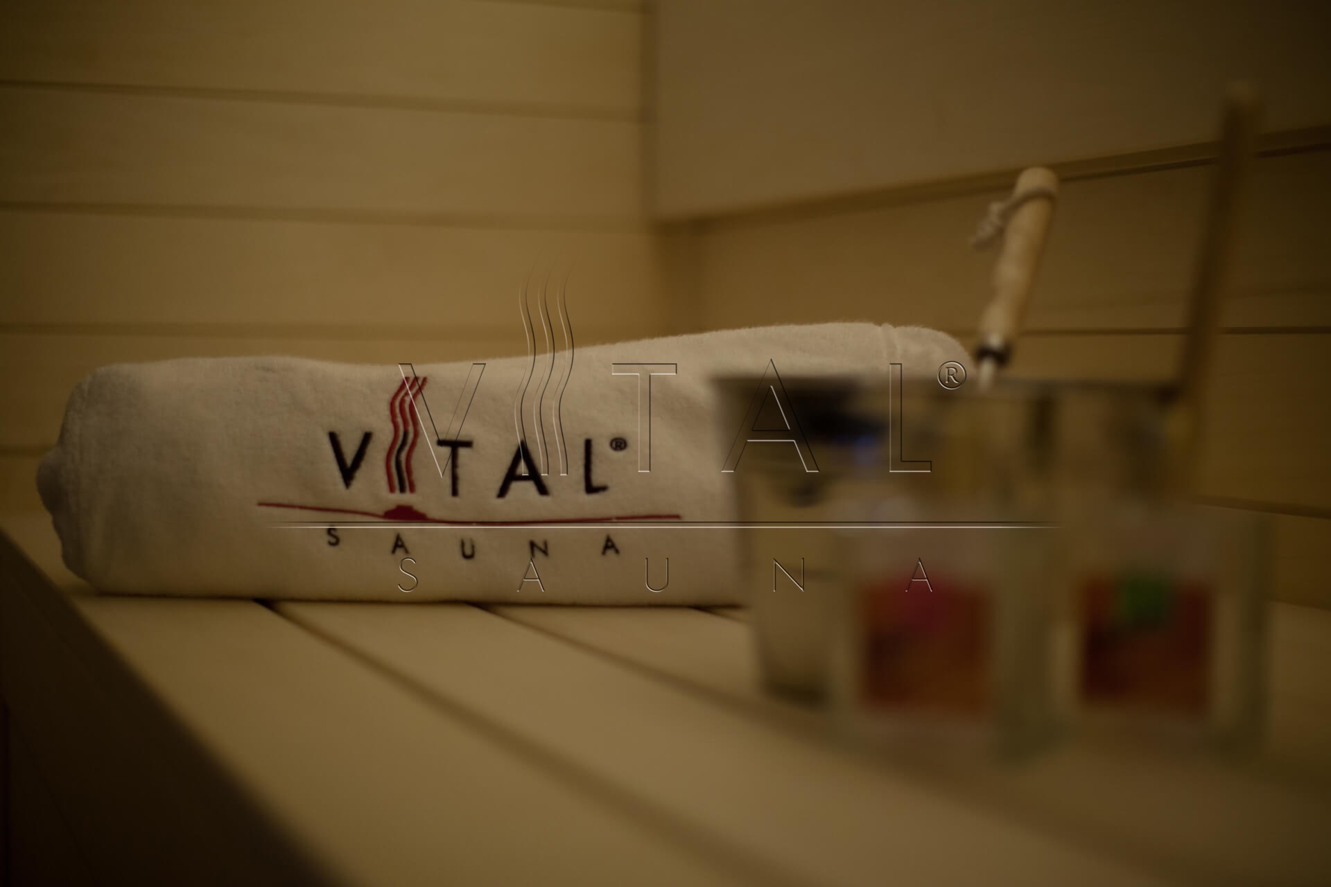 Wnętrze drewnianej kabiny, ręcznik z napisem VITAL SAUNA i wiadro z chochlą na ławce.