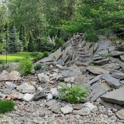 Salon Zieleni - Aranżacja skalniaka z kamiennymi schodkami, roślinami ozdobnymi i żwirowym podłożem. W tle zielony trawnik i drzewa.