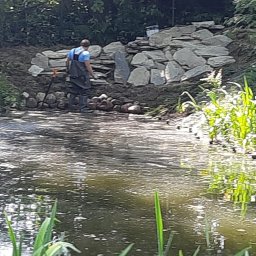 Salon Zieleni - Pracownik w woderach, z łopatą, buduje kaskadę z kamieni nad brzegiem stawu. W tle widoczne drzewa i roślinność wodna.