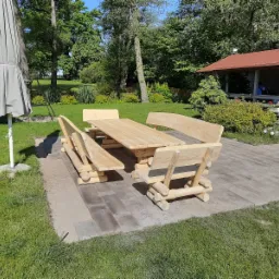 Rustykalny zestaw mebli ogrodowych z surowego drewna na tle zielonego trawnika i zadbanego ogrodu. Widoczny parasol i domek ogrodowy.