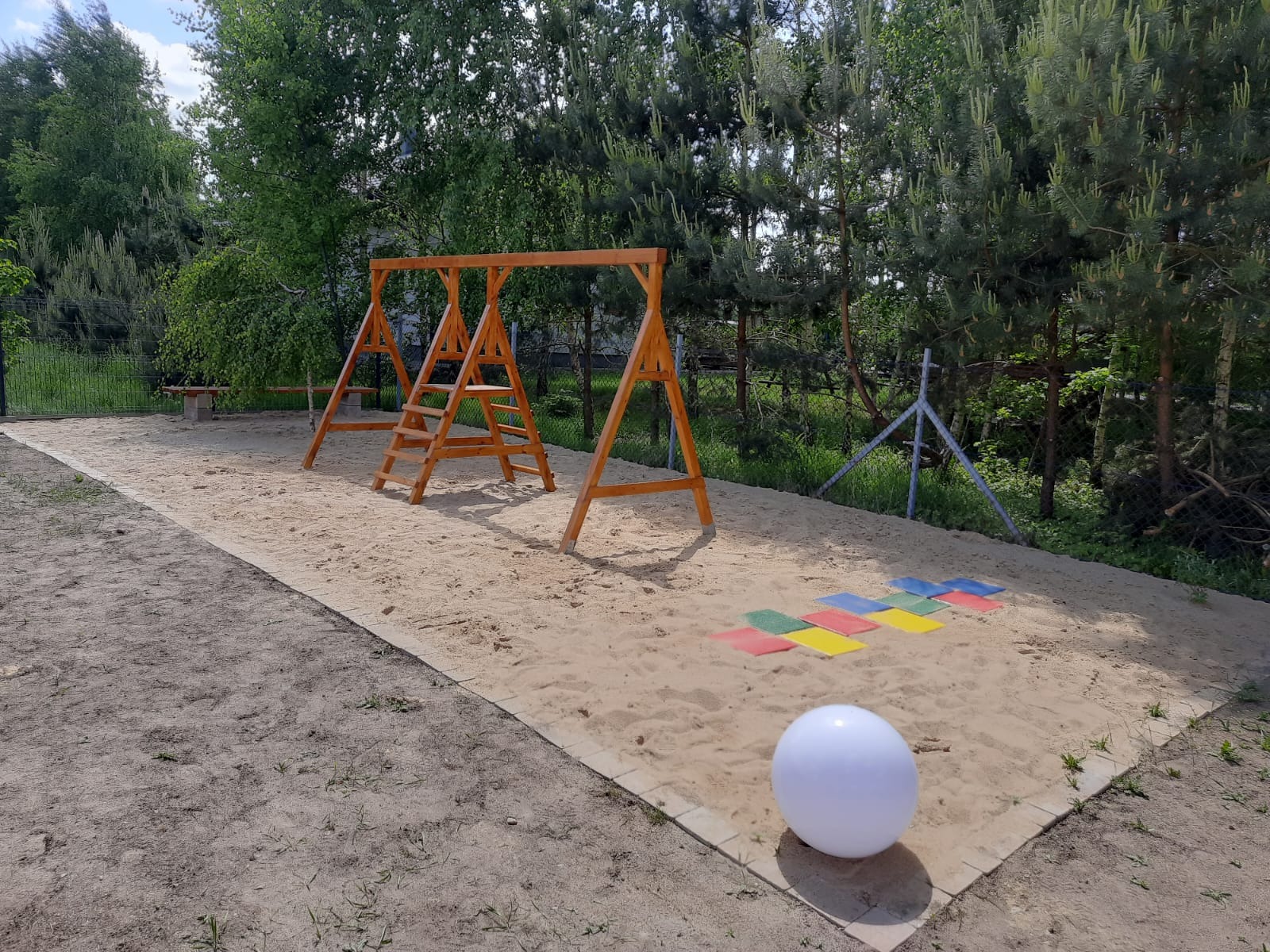 Plac zabaw z piaskiem, drewnianą huśtawką i kolorowymi płytkami do skakania. W tle drzewa i ogrodzenie. Biała kula na pierwszym planie.