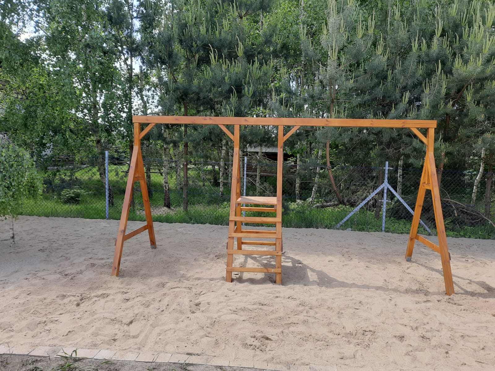 Drewniana konstrukcja huśtawki z drabinką na piaszczystym placu zabaw, w tle drzewa i siatka ogrodzeniowa. Naturalne oświetlenie.