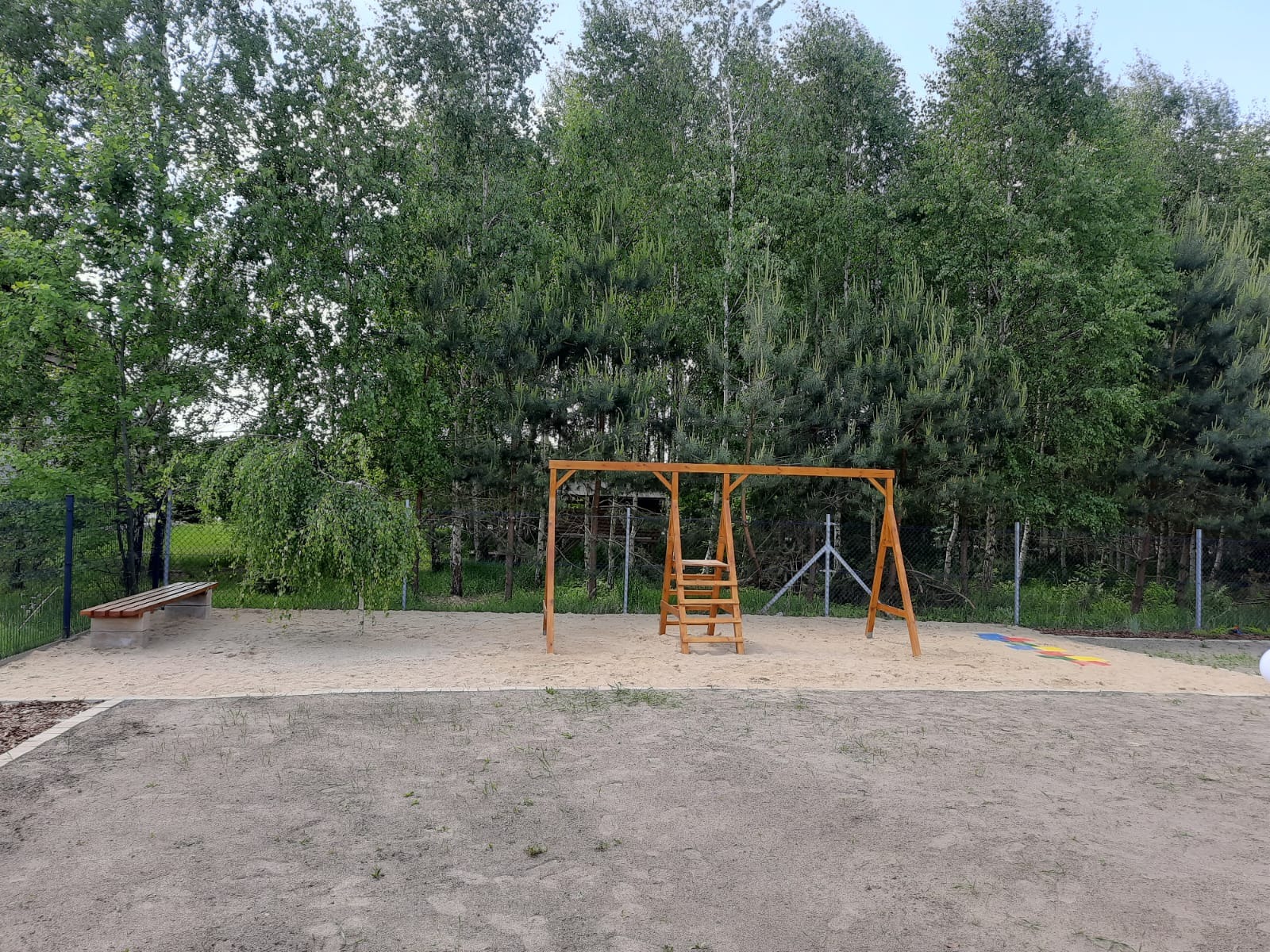 Plac zabaw z piaskiem, drewnianą huśtawką drabinkową, ławką i młodym drzewem płaczącym na tle zieleni i ogrodzenia z siatki.