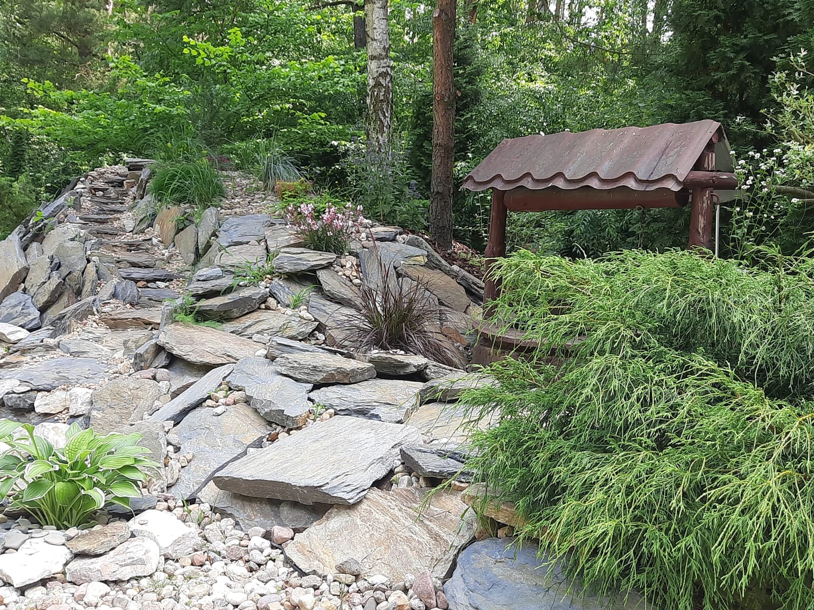 Ogród skalny z kaskadą z płaskich kamieni, zielenią i drewnianą studnią. Ujęcie z poziomu gruntu, podkreślające naturalny charakter aranżacji.