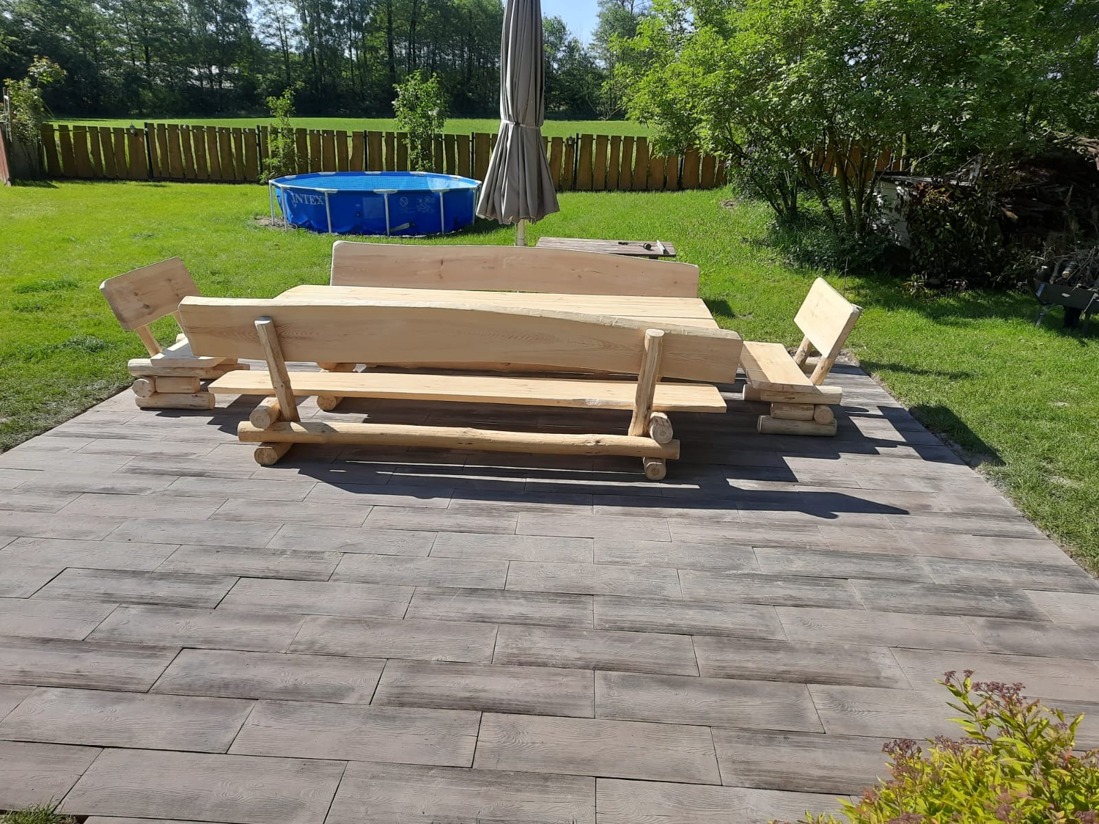 Drewniany stół z ławkami i krzesłami na tarasie z płytek imitujących drewno. W tle trawnik, ogrodzenie i basen.