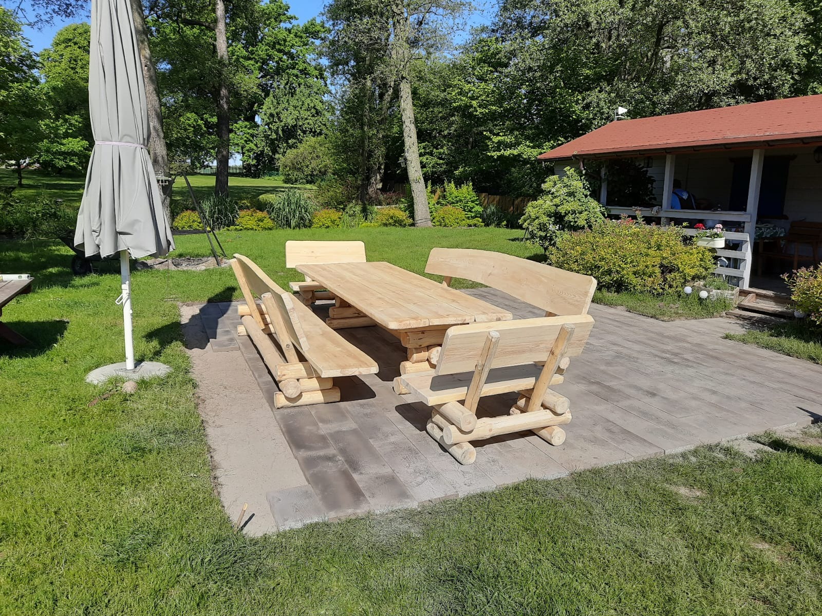 Rustykalny zestaw mebli ogrodowych z surowego drewna na tle zielonego trawnika i zadbanego ogrodu. Widoczny parasol i domek ogrodowy.