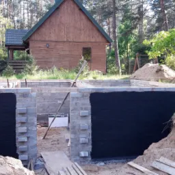 Fundamenty domu w trakcie budowy, zabezpieczone czarną izolacją, na tle drewnianego domu i lasu.
