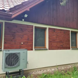 Zewnętrzna jednostka klimatyzacyjna, prawdopodobnie z funkcją grzania, zamontowana na metalowej konstrukcji przy elewacji domu z drewnianą boazerią i trzema oknami.