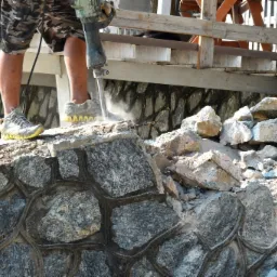 Pracownik w kamuflażowych szortach kruszy kamienny murek młotem pneumatycznym, spod narzędzia wydobywa się pył, w tle widoczna drewniana konstrukcja.