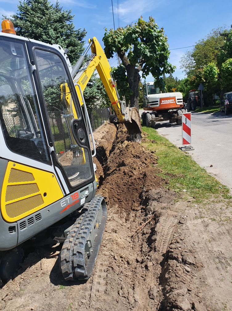 Żółto-szara minikoparka ET18 wykonuje wykop wzdłuż drogi, z widocznymi śladami opon i drugim, większym pojazdem budowlanym w tle.