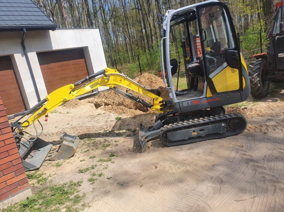 Żółto-szara mini koparka Wacker Neuson ET16 wykonuje prace ziemne na piaszczystym terenie obok budynku z brązową bramą garażową, widoczny fragment czerwonej ściany z cegły i drzewa w tle.