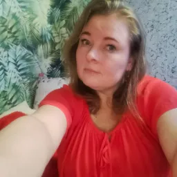 Kobieta w czerwonej bluzce robiąca selfie na tle ściany z wzorem tropikalnych liści i drugiej ściany z fakturą betonu