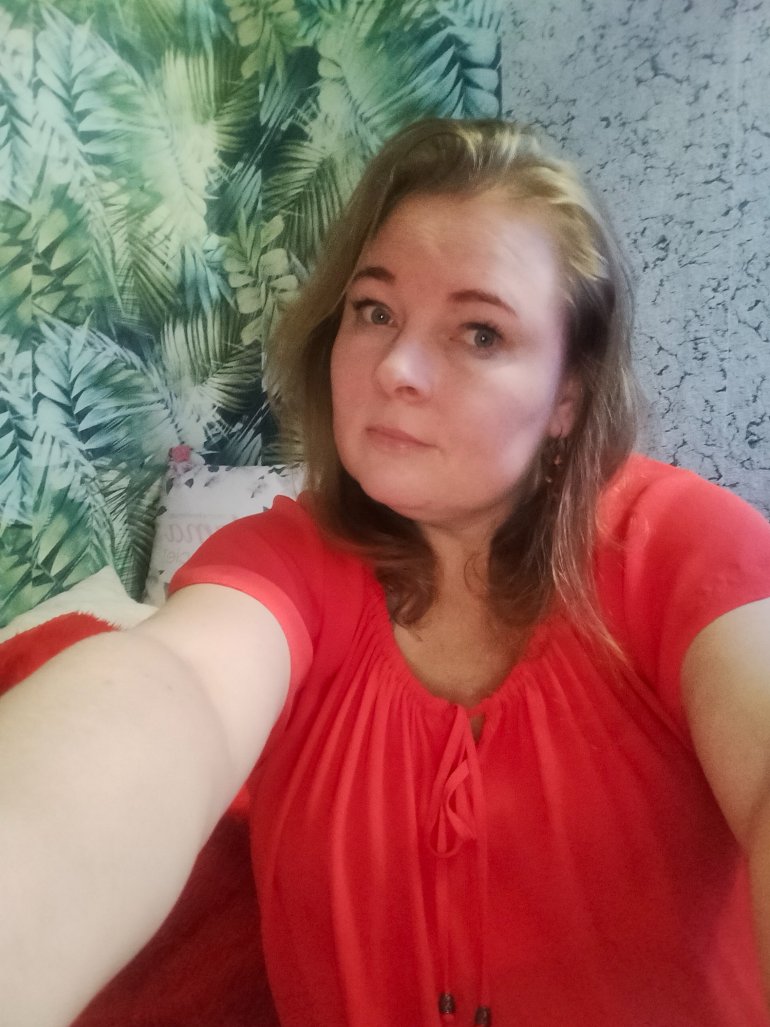 Kobieta w czerwonej bluzce robiąca selfie na tle ściany z wzorem tropikalnych liści i drugiej ściany z fakturą betonu