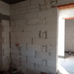 Surowa ściana z betonu komórkowego z wyżłobieniami i poprowadzonymi przewodami elektrycznymi, widoczne puszki instalacyjne i fragment otworu drzwiowego z nadprożem z cegły.