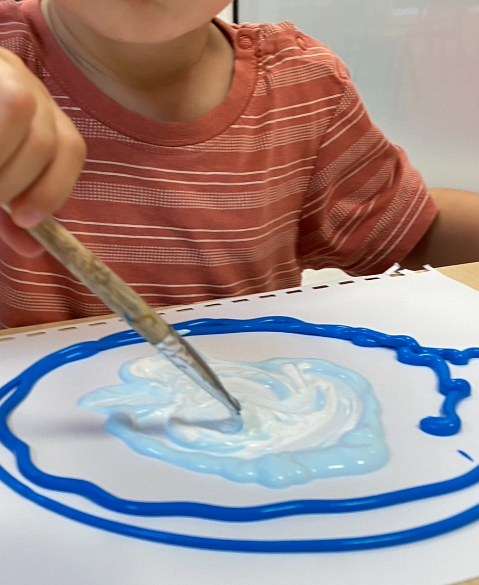 Dziecko w koszulce w paski maluje pędzlem niebieską farbą na białej kartce, tworząc okrągły wzór.