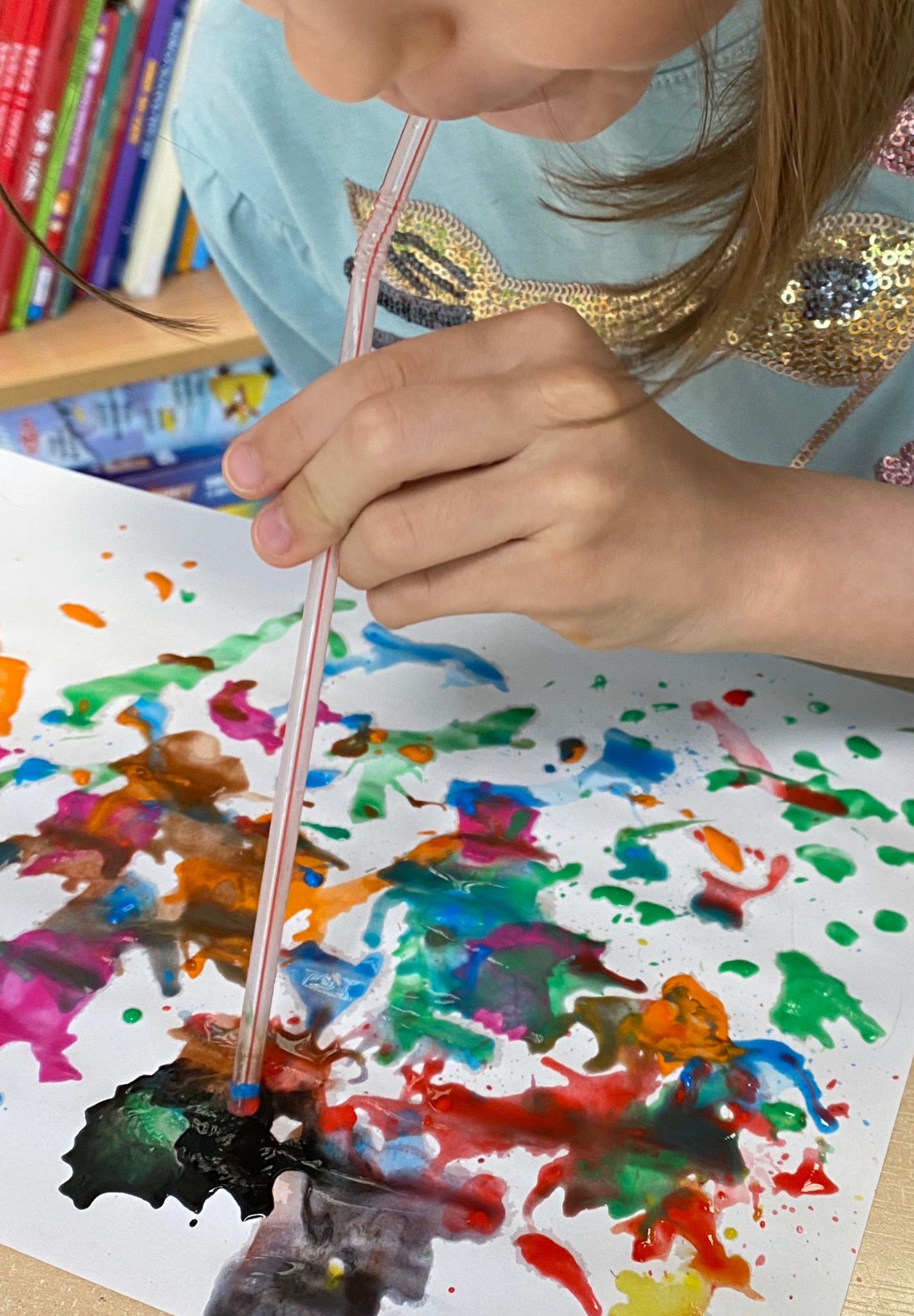 Dziecko dmucha przez słomkę na kleksy farb na białej kartce, tworząc abstrakcyjny obraz. Bluzka dziecka z cekinami w kształcie ryb. Kolorowe książki w tle.