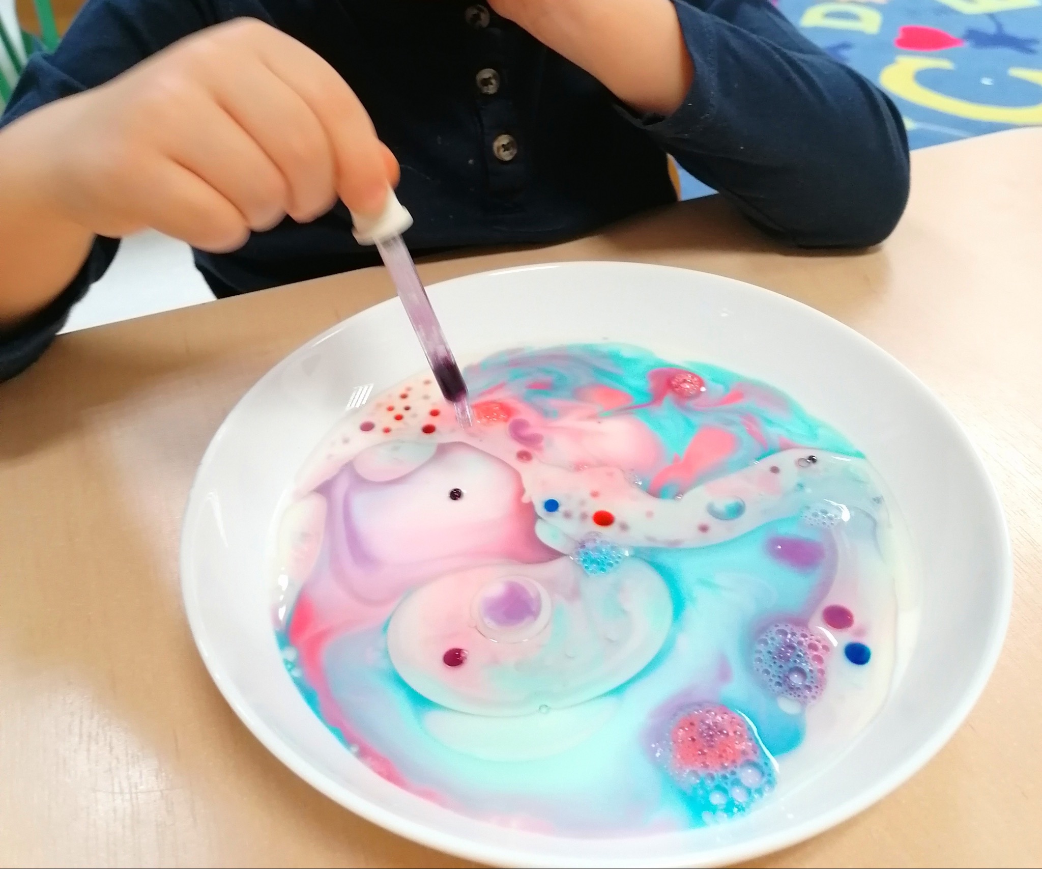 Dziecko dodaje kolorowe krople do mleka w białym talerzu za pomocą pipety, tworząc artystyczne wzory.