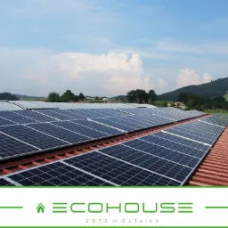 Rząd paneli słonecznych na dachu z czerwonej dachówki, w tle zielone wzgórza i błękitne niebo z białymi chmurami, w dolnej części widoczne logo firmy ECOHOUSE.