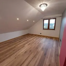 Poddasze z drewnianą podłogą, oknem i żyrandolem w stylu glamour, oświetlone punktowymi lampami sufitowymi.