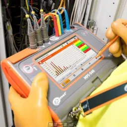 Elektryk w pomarańczowych rękawicach ochronnych używa specjalistycznego miernika Sonel MPI-540 do analizy instalacji elektrycznej. Na ekranie miernika widoczny wykres harmonicznych. W tle widoczne...