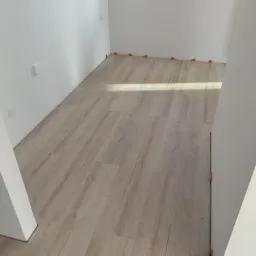 Układanie paneli podłogowych w jasnym, nowym pomieszczeniu z białymi ścianami, widoczne pomarańczowe kliny dystansowe przy ścianach i ościeżnicy.