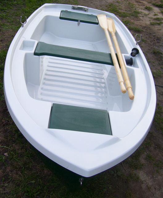 Nowa, biała łódź wiosłowa z zielonymi elementami, widziana z góry, z dwoma drewnianymi wiosłami opartymi o burtę, na tle trawy.