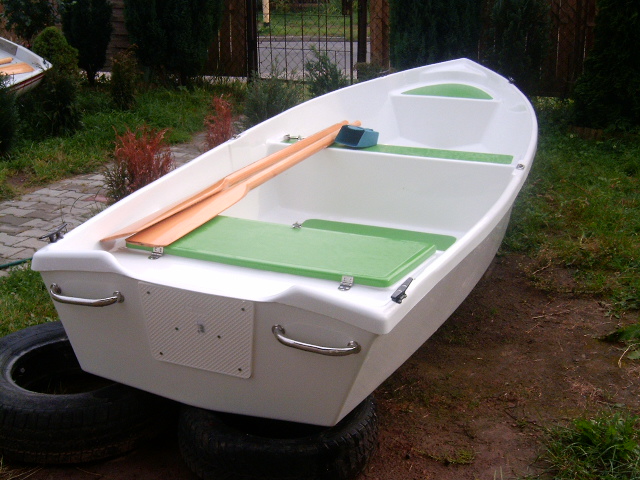 Nowa, biała łódź wiosłowa z zielonymi akcentami, umieszczona na oponach w ogrodzie, wiosła leżące wewnątrz.