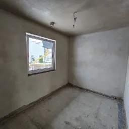 Surowe wnętrze pokoju po tynkowaniu maszynowym; widoczne szare ściany, sufit i podłoga w trakcie prac wykończeniowych, białe okno z widokiem na dom sąsiada.