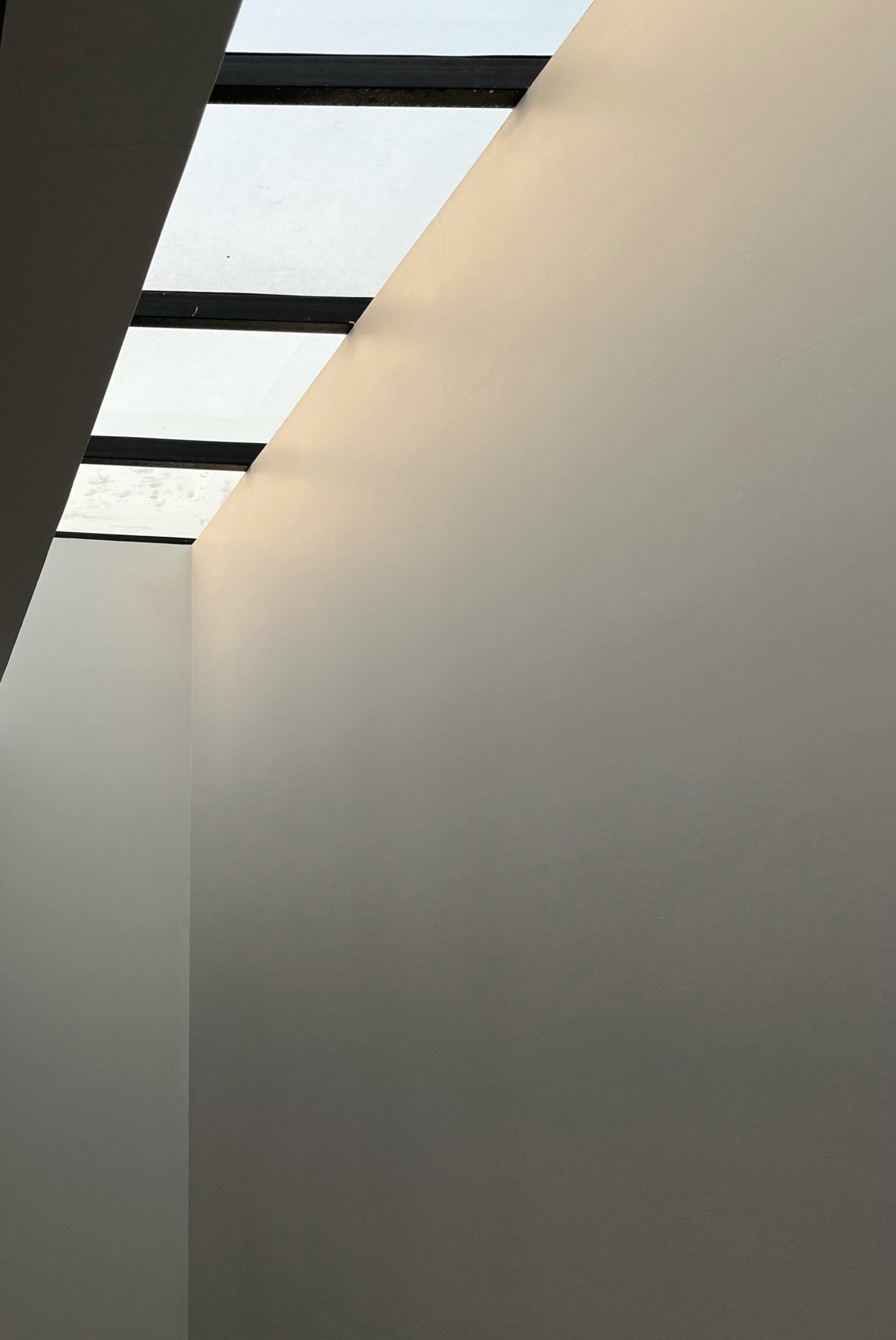 Wnętrze z gładkimi, pomalowanymi na jasny odcień ścianami, oświetlone przez okno dachowe z czarnymi ramami, ukazujące minimalistyczny kąt pomieszczenia.