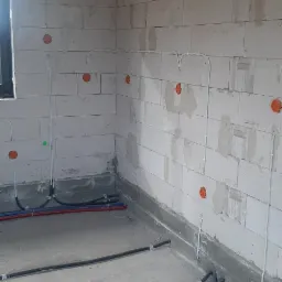 Surowy stan: Wnętrze w trakcie prac, widoczne kable elektryczne w peszlach na podłodze i białe kable prowadzone po ścianach z betonu komórkowego do pomarańczowych puszek instalacyjnych.