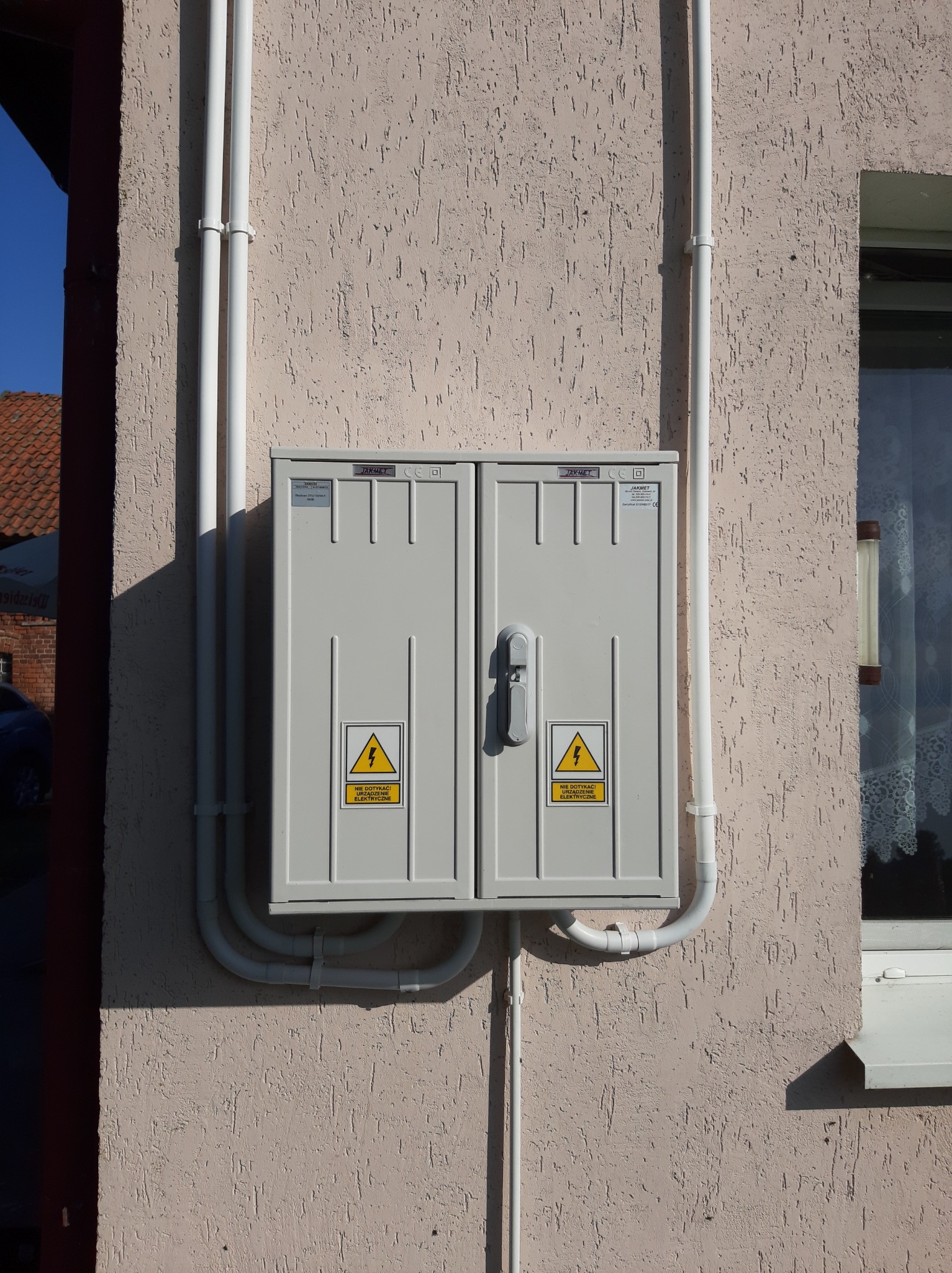 Szara skrzynka elektryczna z ostrzeżeniami na elewacji budynku, z białymi rurami PCV doprowadzającymi kable, widoczne okno z firanką i fragment dachu w tle.