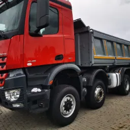 Czerwony samoch&oacute;d ciężarowy Mercedes Arocs 4145 z szarą skrzynią ładunkową typu wywrotka, zaparkowany na kostce brukowej przed budynkiem, widok z przodu i boku.