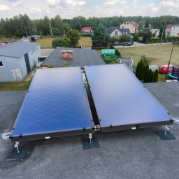 HydroX - Dwa płaskie kolektory słoneczne zamontowane na dachu budynku z widokiem na okoliczną zabudowę i zieleń w słoneczny dzień.