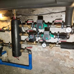 HydroX - Złożona instalacja hydrauliczna z czarnym zbiornikiem buforowym, pompami obiegowymi Wilo, licznymi zaworami z czerwonymi rączkami, manometrami i filtrem TF1 Total, na tle surowej, pomalowanej ściany.