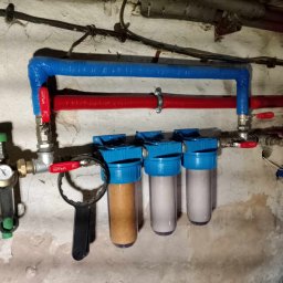 HydroX - Instalacja filtrów wody z manometrem i zaworami, rury w izolacji termicznej w kolorze niebieskim i czerwonym na tle surowej, pomalowanej ściany.