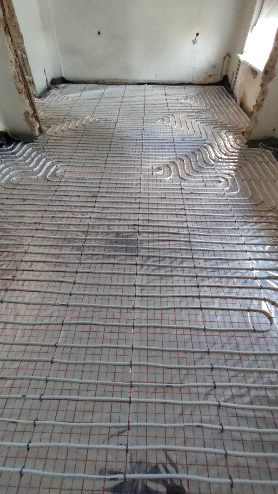 Białe rury ogrzewania podłogowego rozłożone na folii z siatką w surowym wnętrzu, widoczne fragmenty niezabezpieczonych ścian.