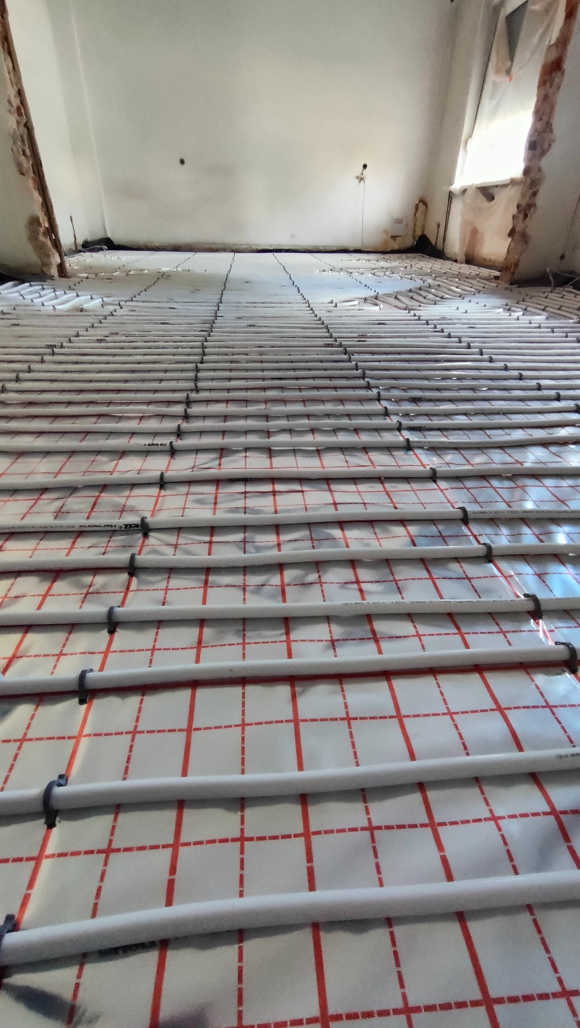 Równomiernie rozłożone białe rury grzewcze zamocowane czarnymi klipsami na folii z czerwoną siatką, przygotowane do wylania posadzki jastrychowej w surowym wnętrzu z fragmentami odsłoniętej cegły.
