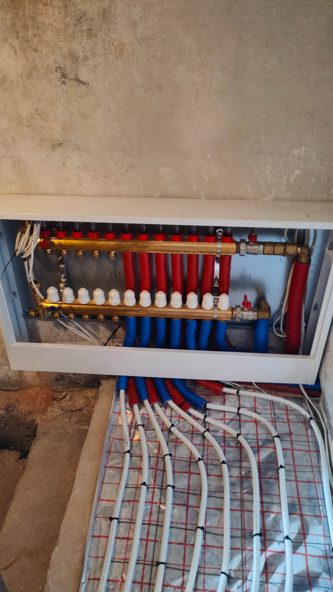 Rozdzielacz ogrzewania podłogowego z czerwono-niebieskimi rurami w białej obudowie, widoczne białe rurki na folii z siatką, surowa ściana w tle.