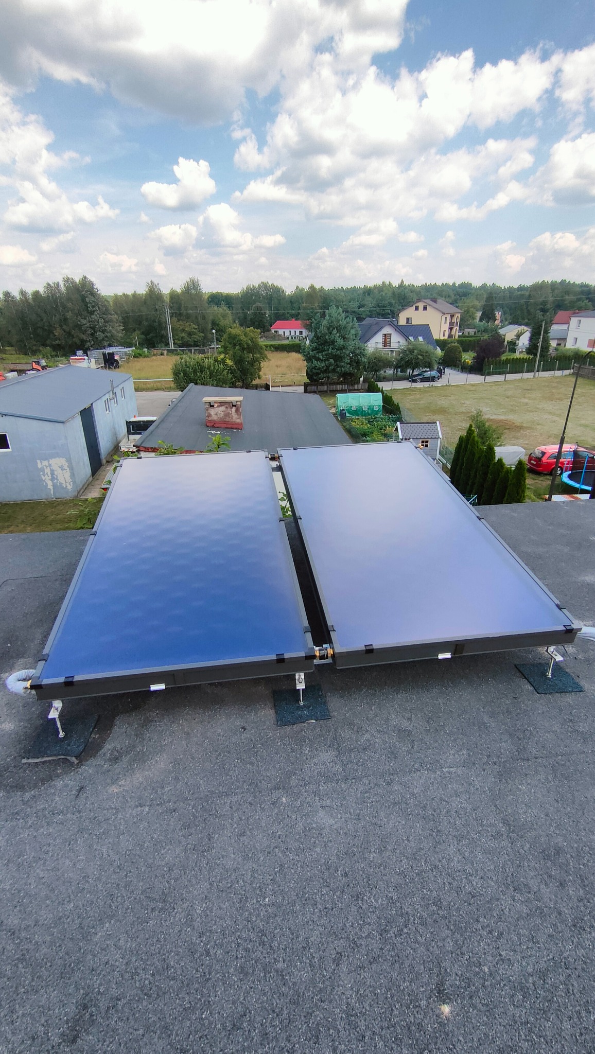 Dwa płaskie kolektory słoneczne zamontowane na dachu budynku z widokiem na okoliczną zabudowę i zieleń w słoneczny dzień.