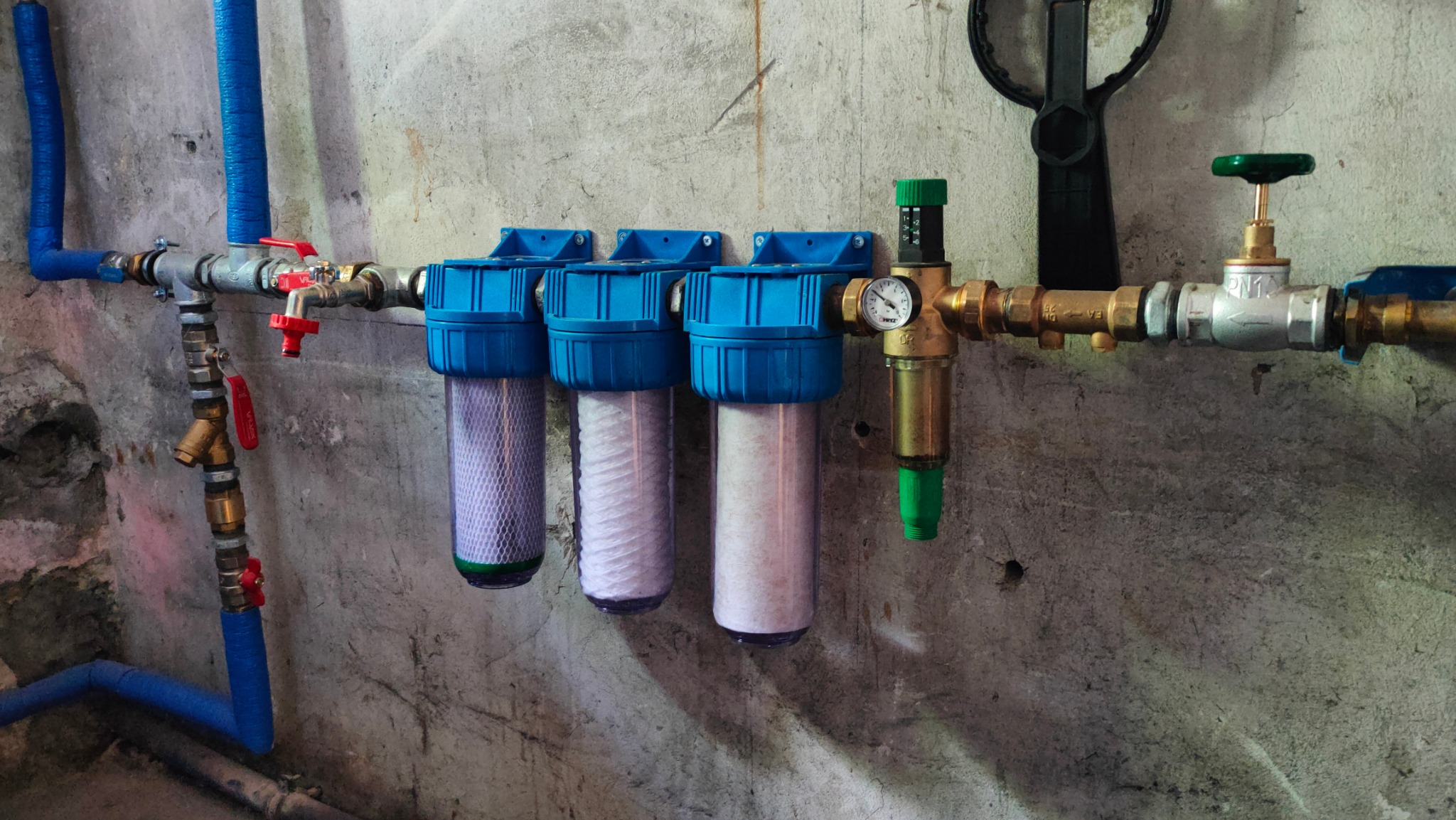 Trzy filtry wody z niebieskimi obudowami zamontowane na ścianie z widocznymi rurami i zaworami, w tym jeden z manometrem.