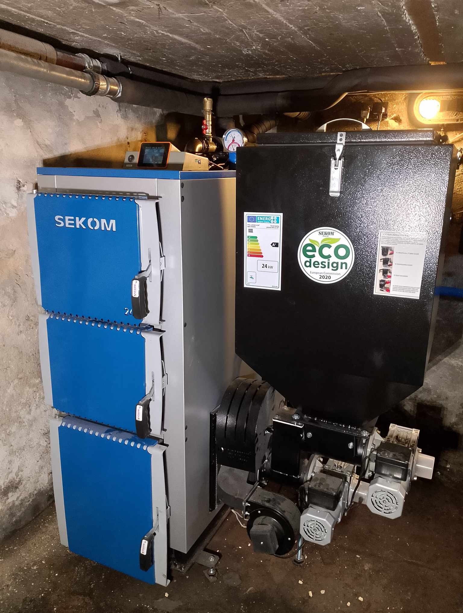 Piec CO Sekom z podajnikiem na ekogroszek, z widoczną etykietą energetyczną i naklejką eco design 2020, zainstalowany w kotłowni z rurami pod sufitem.