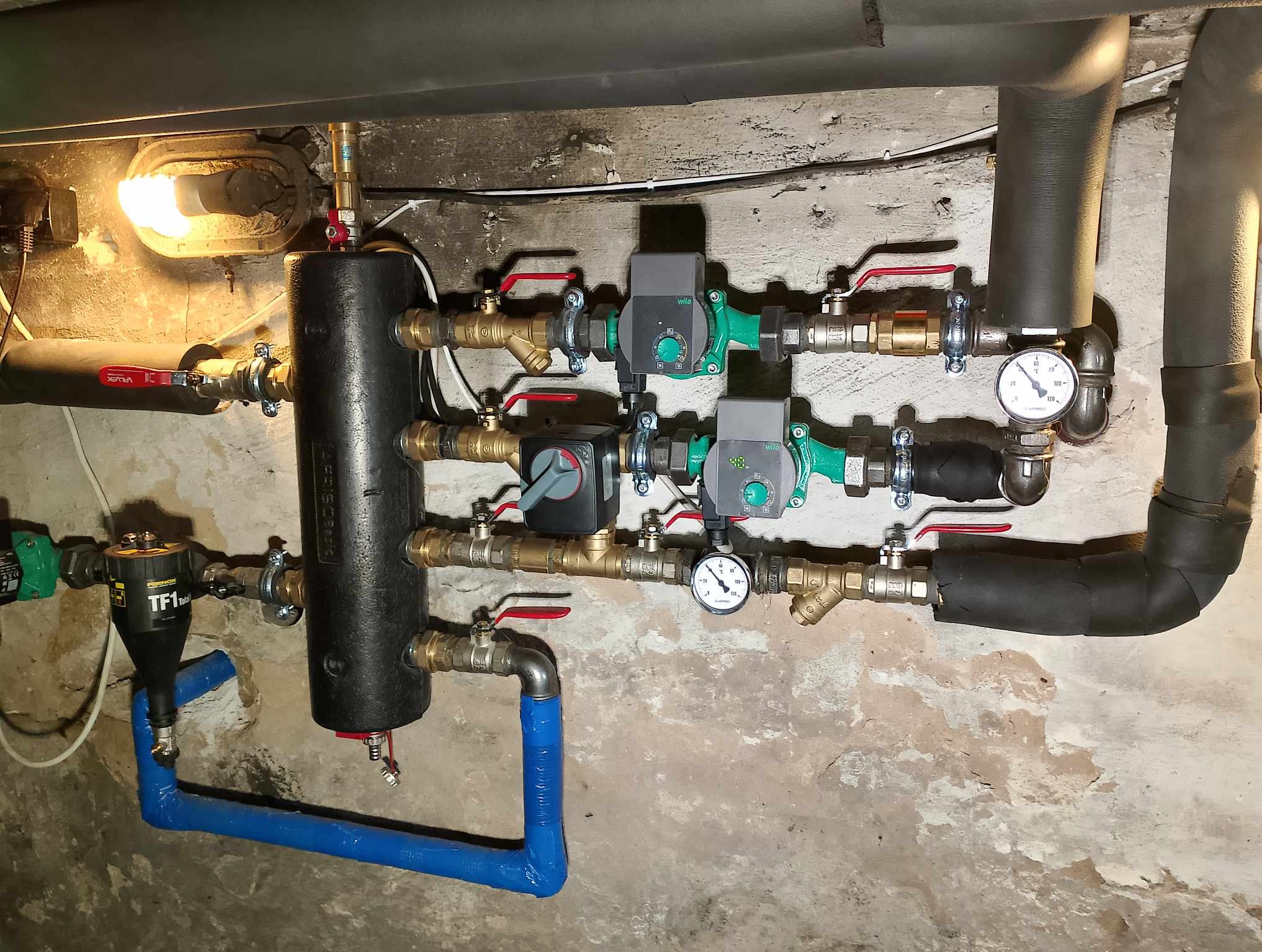 Złożona instalacja hydrauliczna z czarnym zbiornikiem buforowym, pompami obiegowymi Wilo, licznymi zaworami z czerwonymi rączkami, manometrami i filtrem TF1 Total, na tle surowej, pomalowanej ściany.