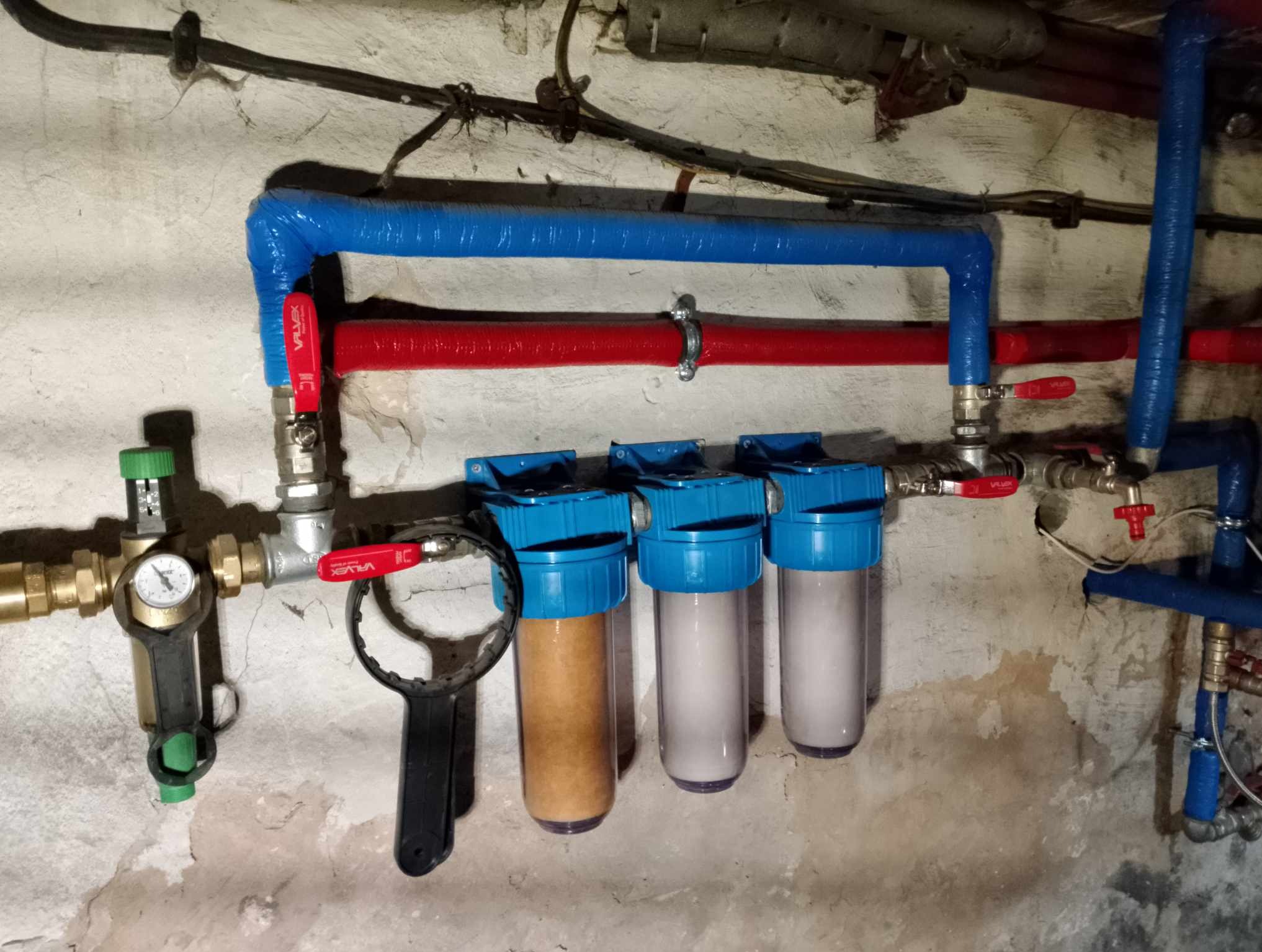 Instalacja filtrów wody z manometrem i zaworami, rury w izolacji termicznej w kolorze niebieskim i czerwonym na tle surowej, pomalowanej ściany.