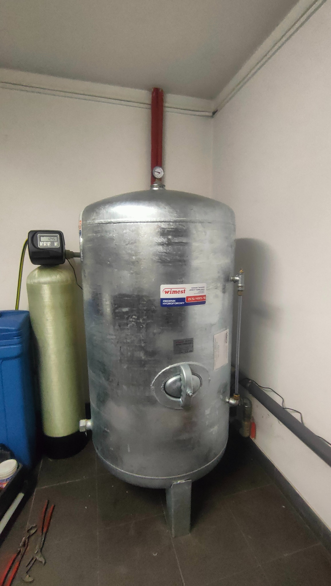 Srebrny zbiornik hydroforowy marki Wimest w kotłowni, obok zielony filtr wody, widoczne narzędzia na podłodze.