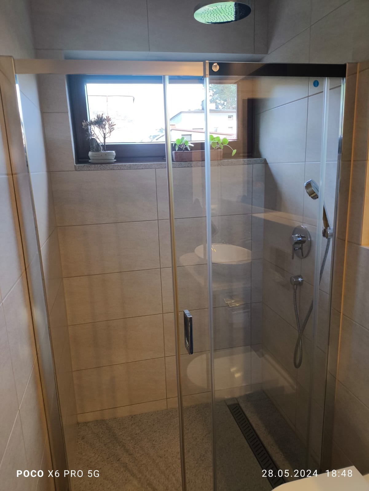 Kabina prysznicowa z przesuwnymi drzwiami, jasne płytki na ścianach, okno z roślinami, deszczownica sufitowa i odpływ liniowy w podłodze.