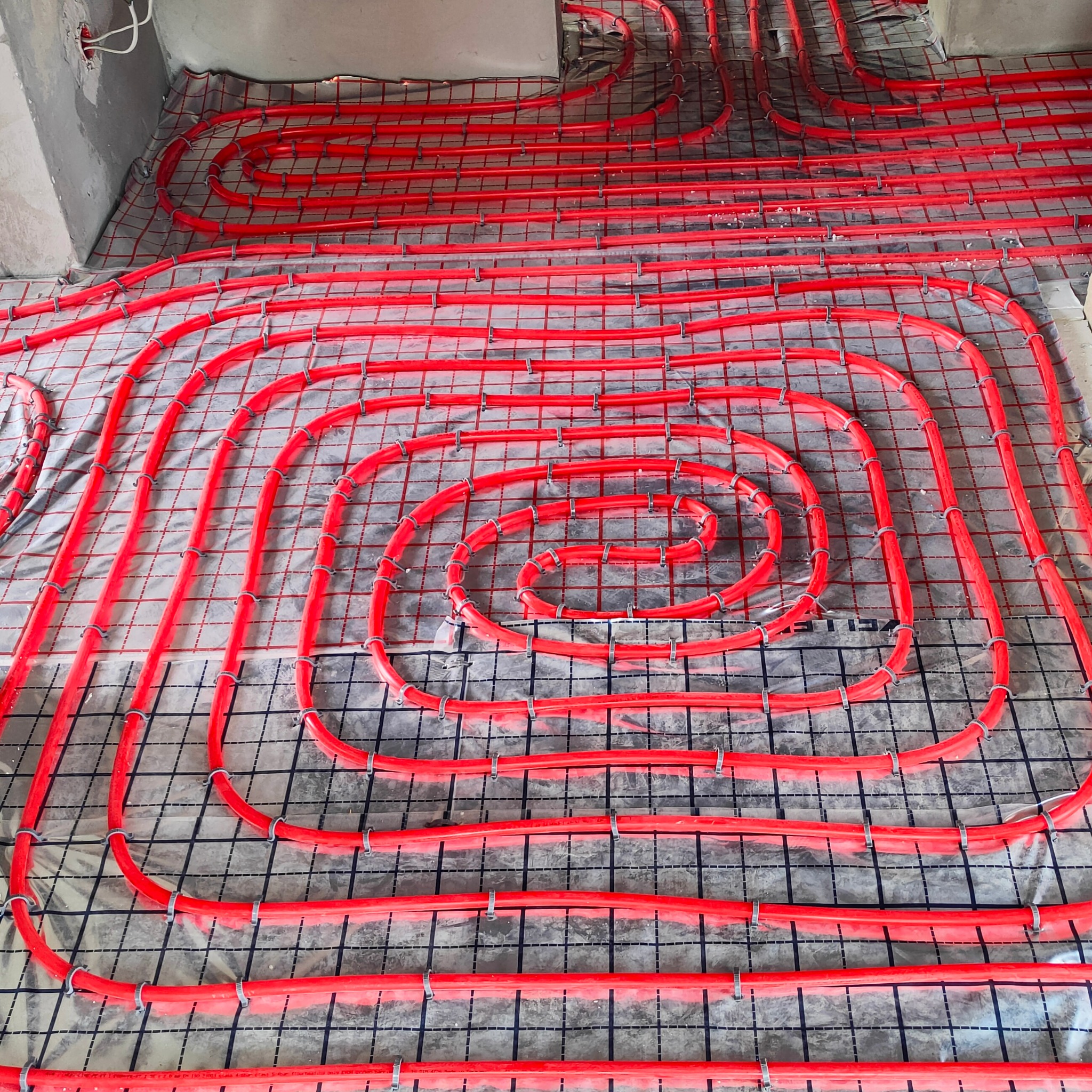 Czerwone rury ogrzewania podłogowego ułożone spiralnie na folii z siatką, przymocowane plastikowymi klipsami, widoczne kable w ścianie.