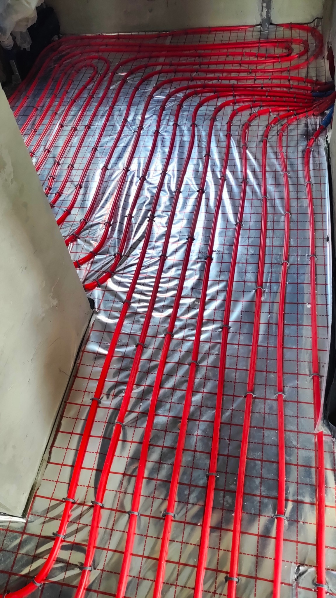 Czerwone rury ogrzewania podłogowego ułożone na srebrnej folii z siatką w trakcie montażu w surowym wnętrzu.