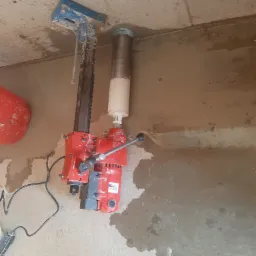 Wiertnica diamentowa, czerwona, przymocowana do sufitu, wierci otwór w betonowym stropie. Widoczne ślady wody i pyłu po wierceniu.