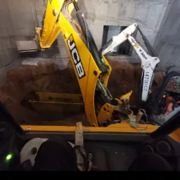 Widok z kabiny żółtej koparki JCB na wykop w betonowym pomieszczeniu, w tle widoczna biała koparka Bobcat.